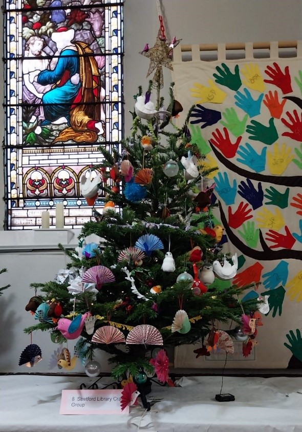 Stretford Library Craft Group's Christmas tree decorated with a variety of handcrafted birds, animals, fans and baubles.  It is displayed in front of a Christian stained glass window on the left-hand side and on the right-hand side, a backcloth of hand shapes painted in a rainbow of colours forming the leaves on the branches of a tree. 