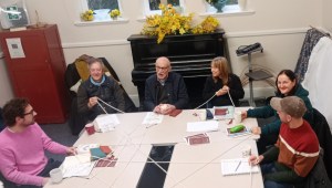 Six members of the group, four men and two women of different ages, seated at one end of the large table, each holding a piece of the web created as part of the exercise.  The web stretches into the foreground, showing there are other members of the group also holding it, not included in the photograph. 