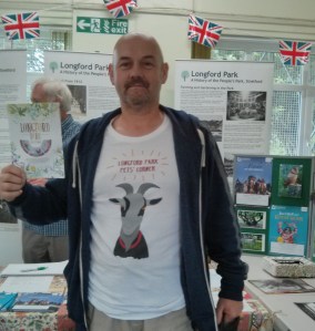 A photograph of Nigel wearing a Pet's Corner tee shirt on which there is a head and shoulders drawing of the pet goat. He is holding a copy of the park information leaflet and map. Behind him is a display telling the history of the Longford Park with photographs, and Union Jack bunting.