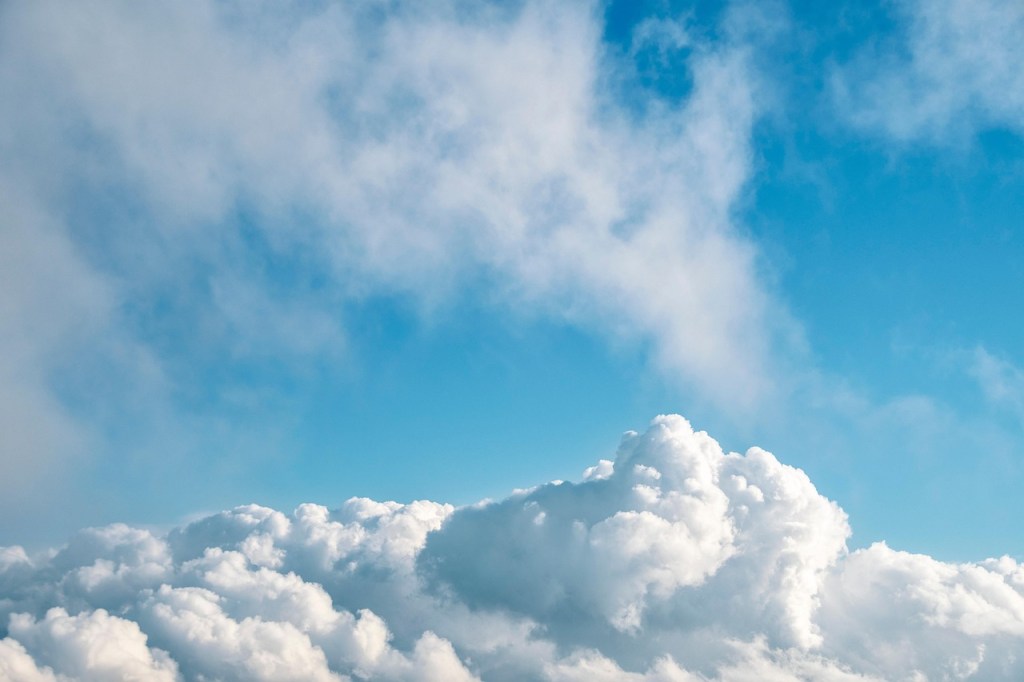 A view of soft, billowing white clouds rising into a bright blue sky. Light wisps of cloud drift across the upper part of the image, giving a sense of openness, calm, and gentle movement.