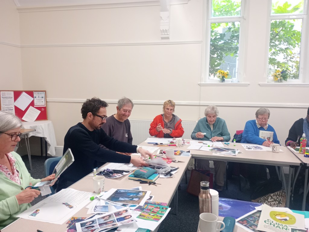 A group of adults engaged in a collage-making activity, cutting and arranging images at tables in a bright room with windows and greenery visible in the background.