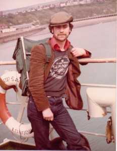 A photograph of Tony as a young man, taken on board a ferry boat, wearing a flat cap, red short, Guinness sweat shirt, brown corduroy jacket and blue jeans with a rucksack on his back. 