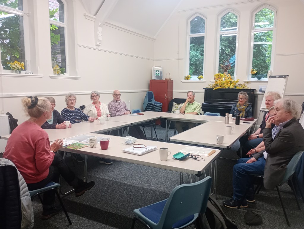 A group of people sitting around a table engaged in a lively discussion during a session on Deaf education, with a presenter in front sharing insights. The room is well-lit with large windows and decorations.