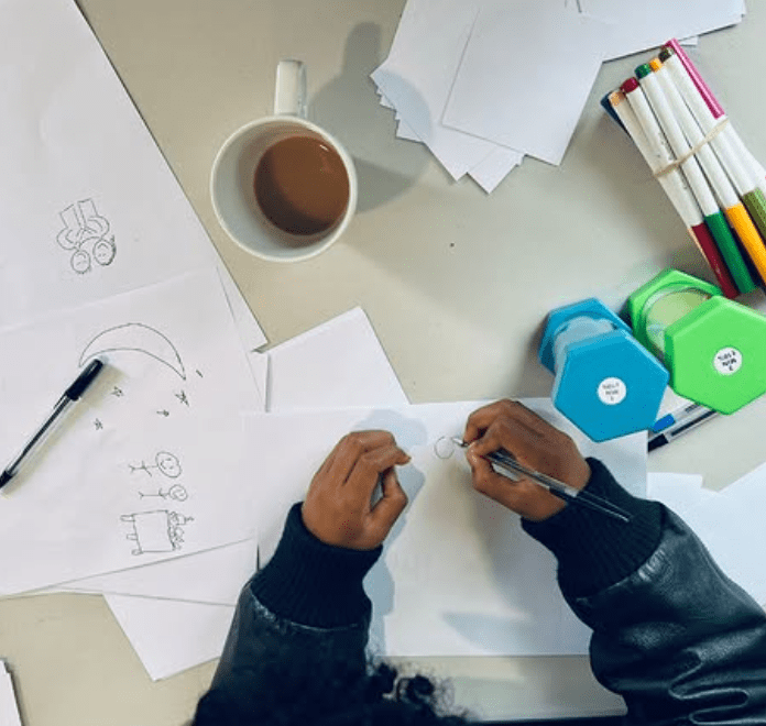An overhead view of hands drawing on paper with a pen, surrounded by a cup of coffee, colorful markers, and blank sheets of paper.