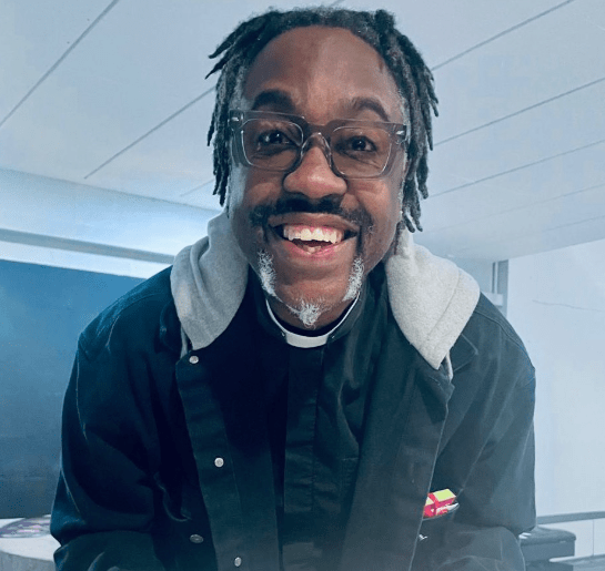 Smiling priest wearing glasses and a casual jacket, welcoming attendees at a community event.