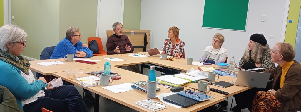 Image: A group of participants engaged in a discussion during a writing workshop at Chorlton Library, with various materials and laptops on the table.