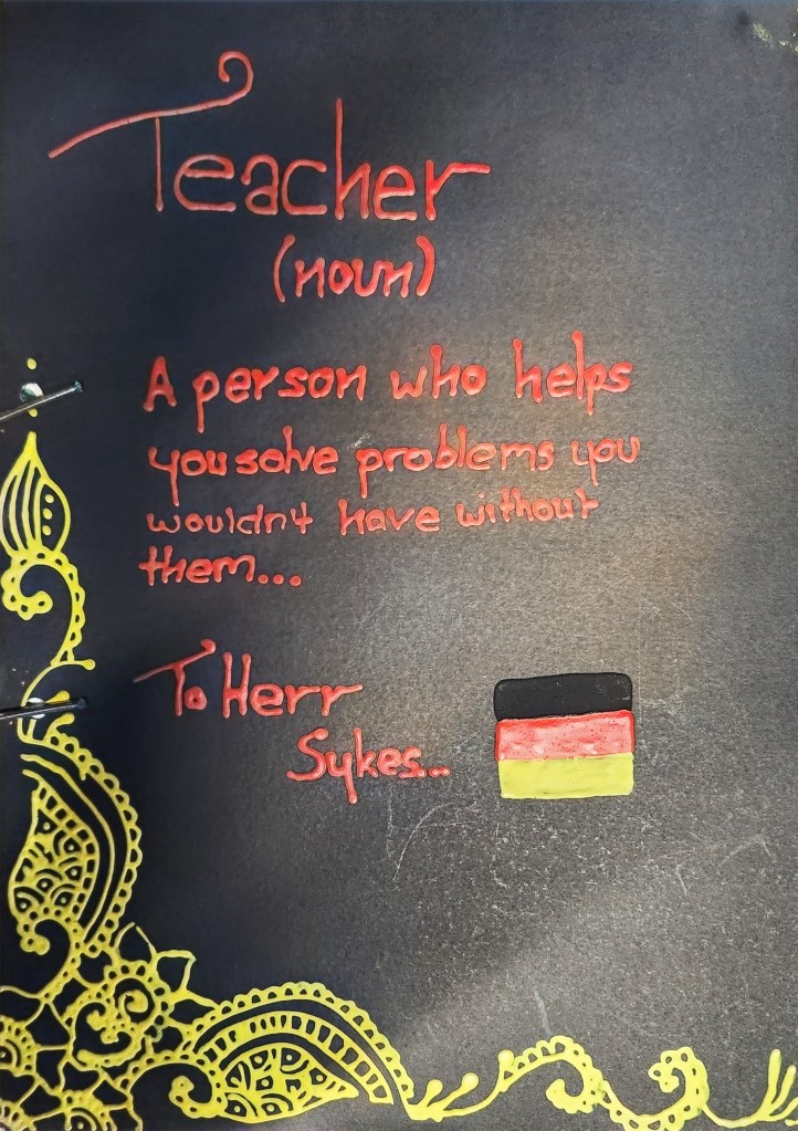 Image: The text in the form of a dictionary entry is written in red fancy writing on a black background with a yellow lace embellishment on the bottom left hand corner. It says "Teacher (noun) A person who helps you solve problems you wouldn't have without them..." and is addressed To Herr Sykes next to a German flag with black, red and yellow horizontal stripes.