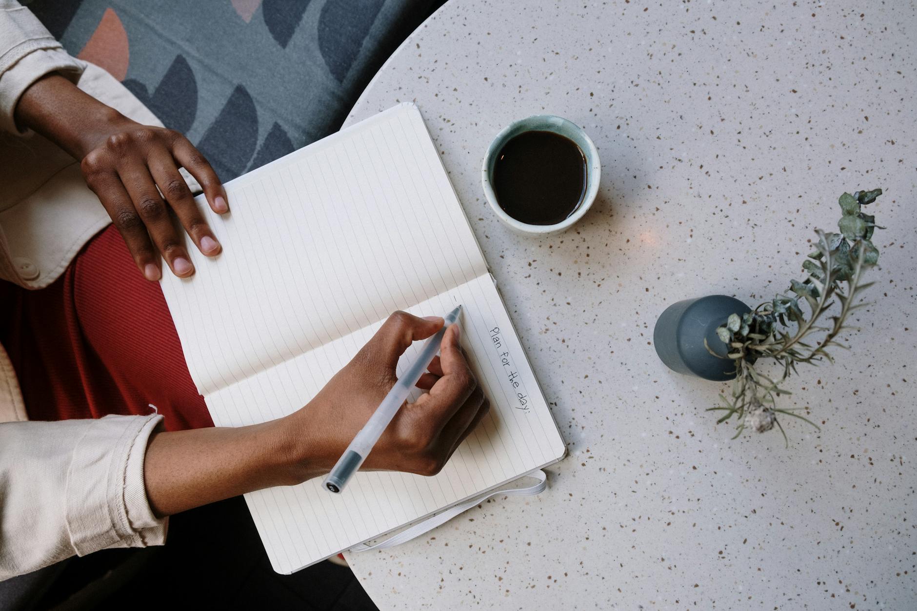 Image: a photo from above of hands writing in a notebook which is resting on a table alongside a cup of coffee.