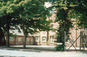 Image: a photograph of the red brick school building and its playground, surrounded by trees.