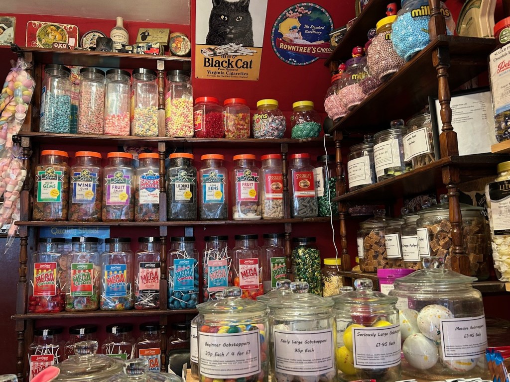 Image: A view of the interior of the sweet shop with rows of jars of sweets on the wooden shelves.  on the counter are jars of gob-stoppers in increasing sizes. 