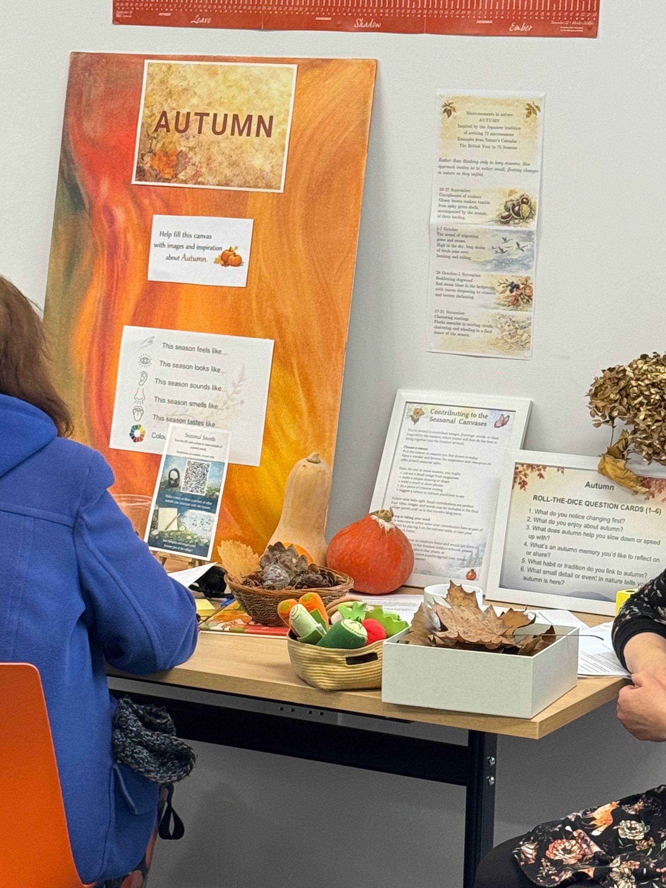 A table setup for an autumn-themed workshop, featuring a colorful canvas labeled 'AUTUMN,' various decorations, and informational sheets about the season. Participants are seen engaging with the materials.