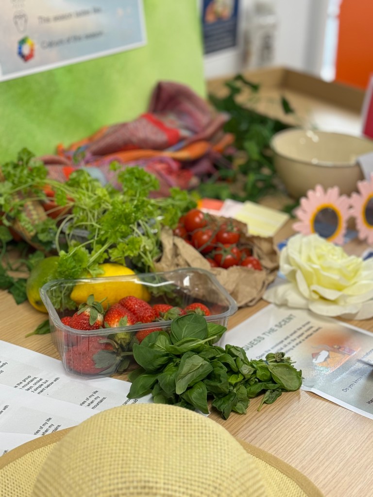 A colorful display of fresh fruits and vegetables on a wooden surface, including strawberries, tomatoes, lemons, and basil, with a decorative cloth and bowls in the background.