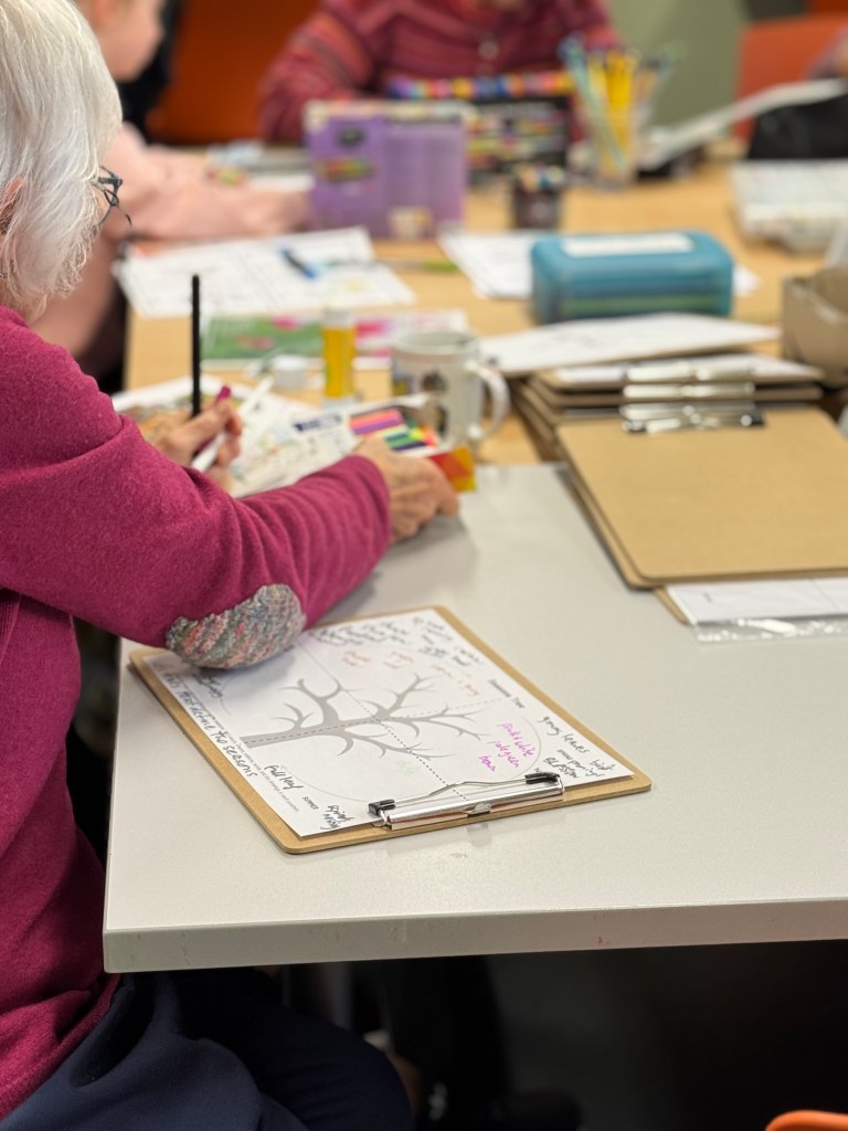 An elderly woman wearing a pink sweater participates in a creative activity, using colorful markers to write on a notepad. Various art supplies and paper are scattered across a table, with other people engaged in similar activities in the background.
