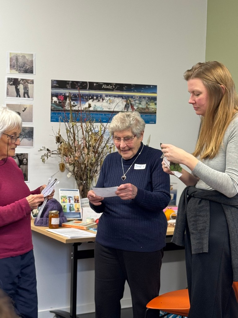 Three women engaged in a discussion, two holding papers with notes. The background features a display of photographs and decorated branches.
