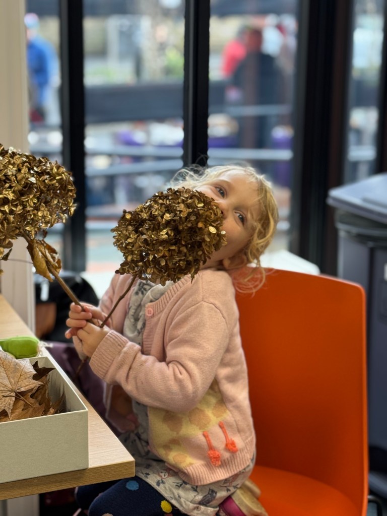 A child sitting at a table holding a decorative flower made of dried leaves, partially hiding her face, with a box of more leaves nearby.