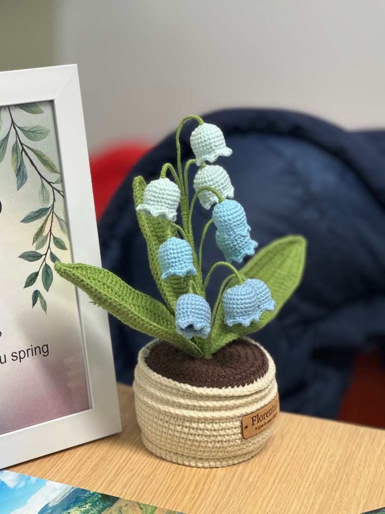 A knitted flower arrangement featuring blue and green bell-shaped flowers in a textured pot, placed on a wooden table with a decorative frame in the background.
