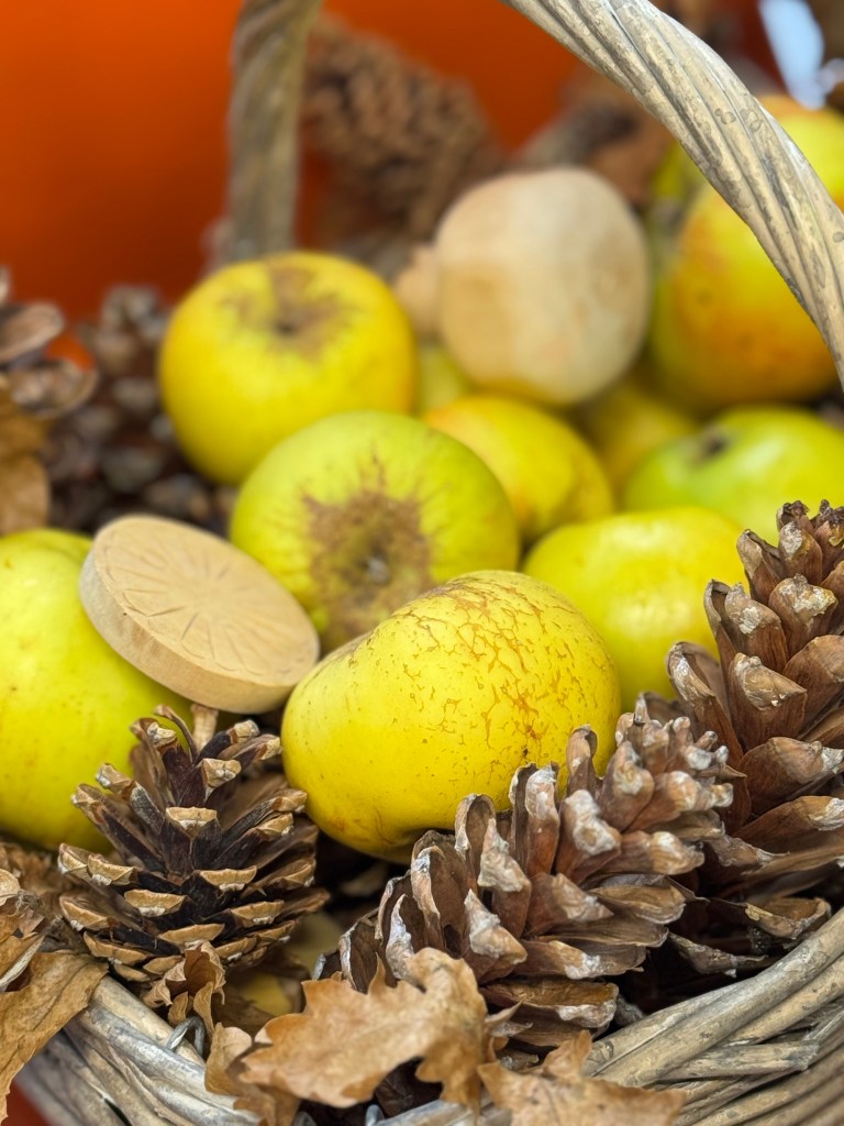 A wicker basket filled with yellow apples, pine cones, and dried leaves, set against a warm orange background.
