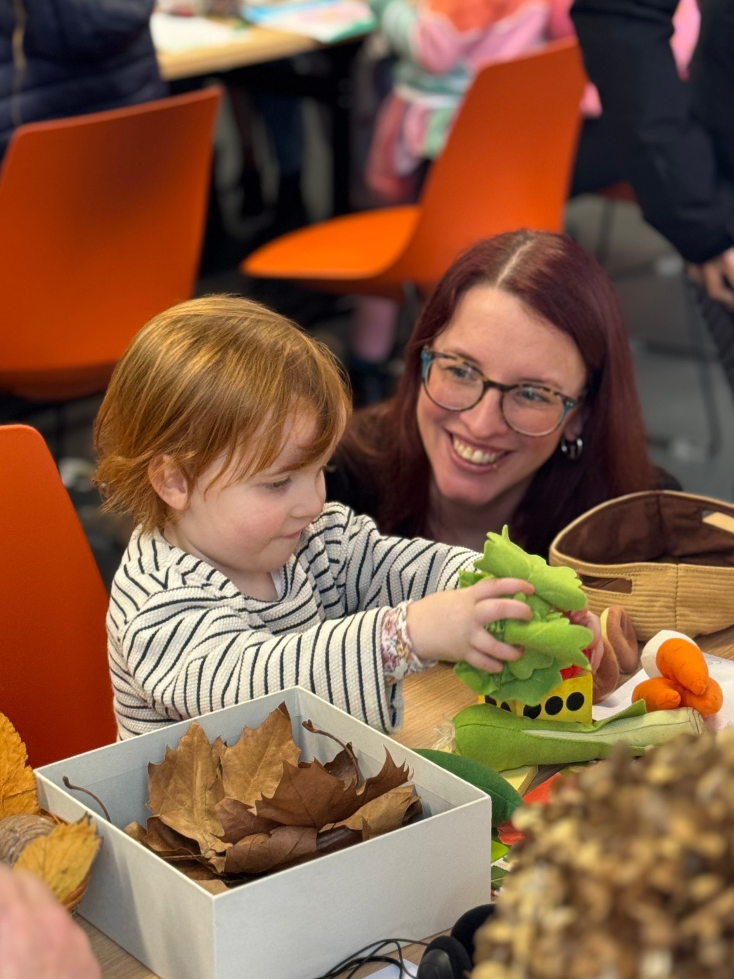 A young child playing with colorful toy vegetables while an adult smiles in the background. Several children are engaged in activities at tables nearby, with autumn leaves in a box on the table.