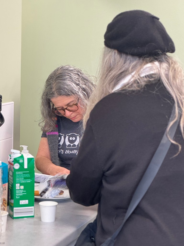 A woman with curly gray hair and glasses is seated at a table, focusing on an item in front of her. She wears a gray shirt with a playful design. In the foreground, another person with long gray hair, wearing a black beret and a black sweater, is partially visible, facing away from the camera. The table is adorned with food items and beverages.