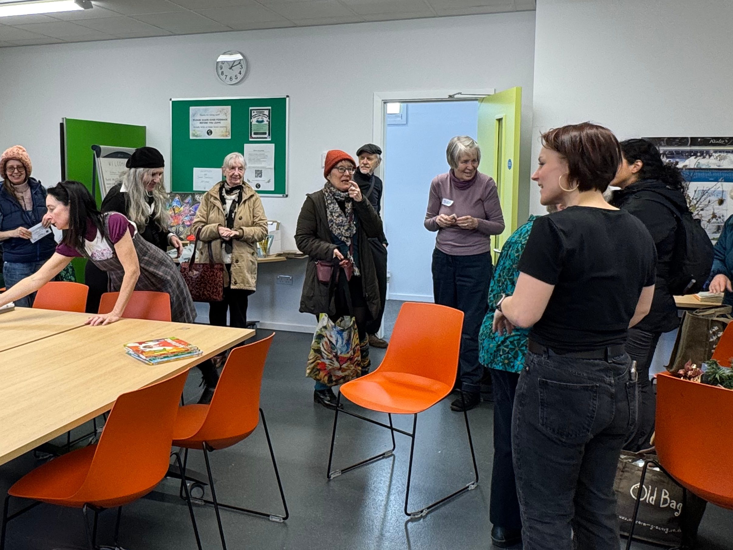 A group of women gathered in a community room with tables and orange chairs, engaging in conversation and activities.