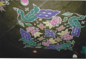 A square Rangoli pattern featuring green and purple peacocks on each of the four corners, each holding two leaves in their beaks. At the centre are floral and geometric patterns in pink, orange and yellow, with a purple flower at the centre.