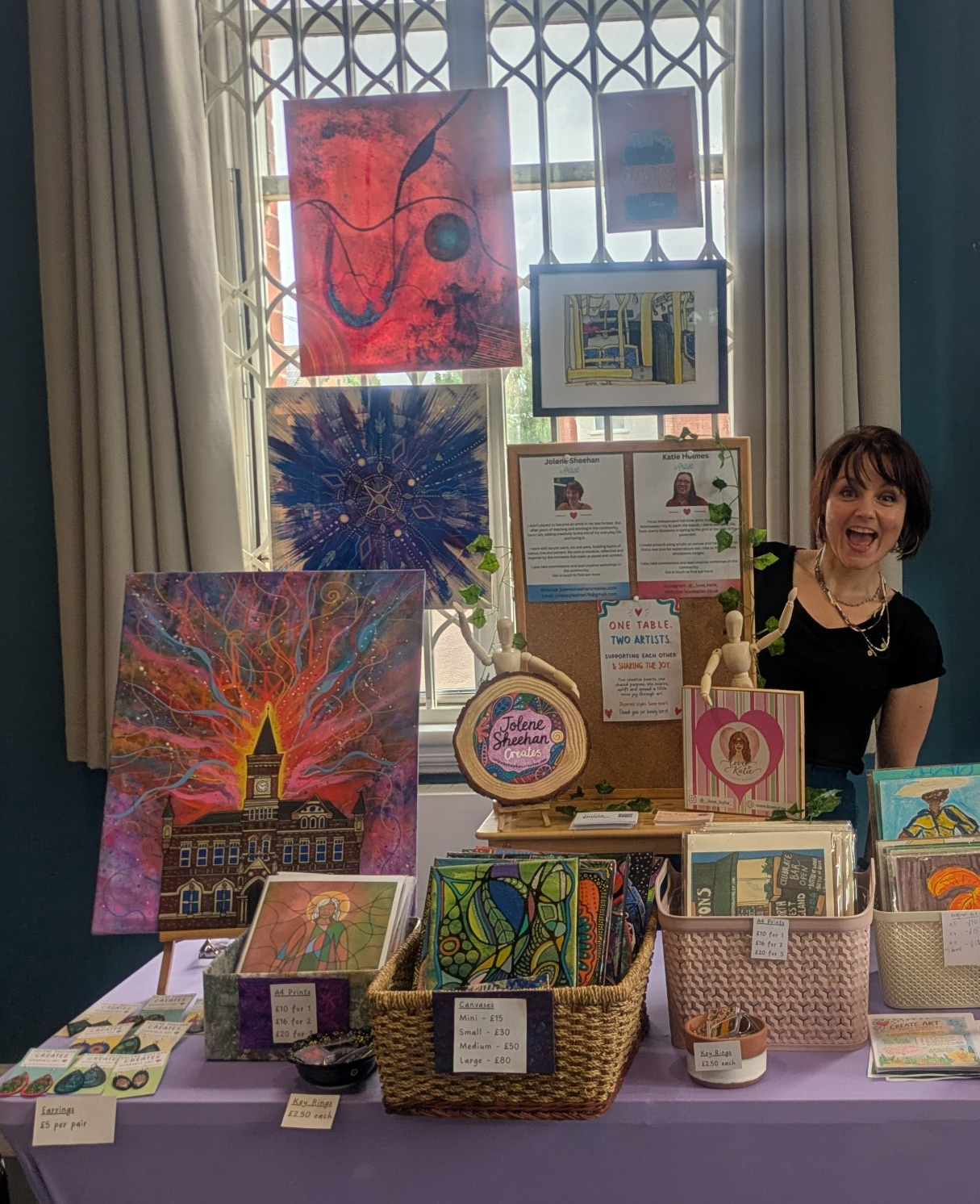 Jolene is peeping out behind a board displaying several brightly coloured paintings. In front of her on a table are displays of her smaller paintings and cards.