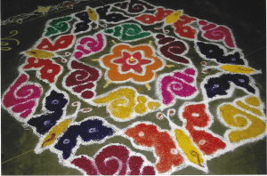 A hexagonal Rangoli pattern on a pavement featuring alternative orange and purple butterflies on each side, with pink, green and yellow snail shells on each corner. There is an orange and red flower in the centre with a lighted candle in the middle.