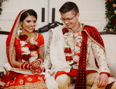 A young Hindu bride and groom. She is wearing a red veil trimmed with gold lace, a red dress and red, black and white bangles. He is wearing a white jacket with a red cloak trimmed with gold. They are both wearing garlands of red and white roses, and both have red bindis on their foreheads.