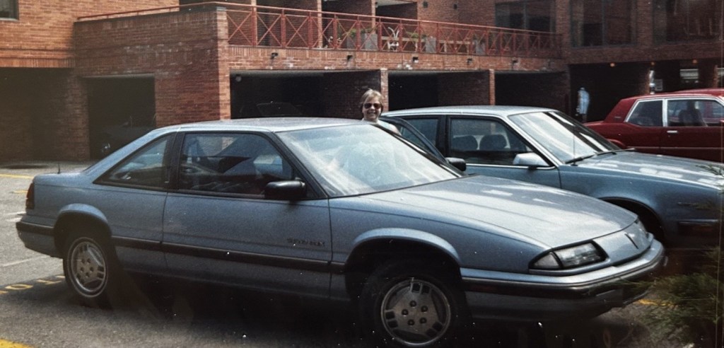 Image description: Margaret with her head just visible over the top of the car, at the driver's door of the Pontiac.
