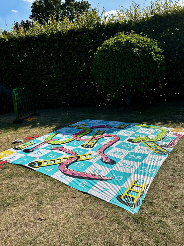 A photograph of the giant snakes and ladders board stretched out and pinned down on a lawn besides a large hedge.  It is a simplified board, numbered 1 to 80, with six pink or green snakes and seven ladders. 