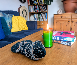 Image description: a photograph of a cosy living room, with a welcoming cushioned sofa in front of which is a coffee table with a lit candle, a pile of books and a woollen hat.