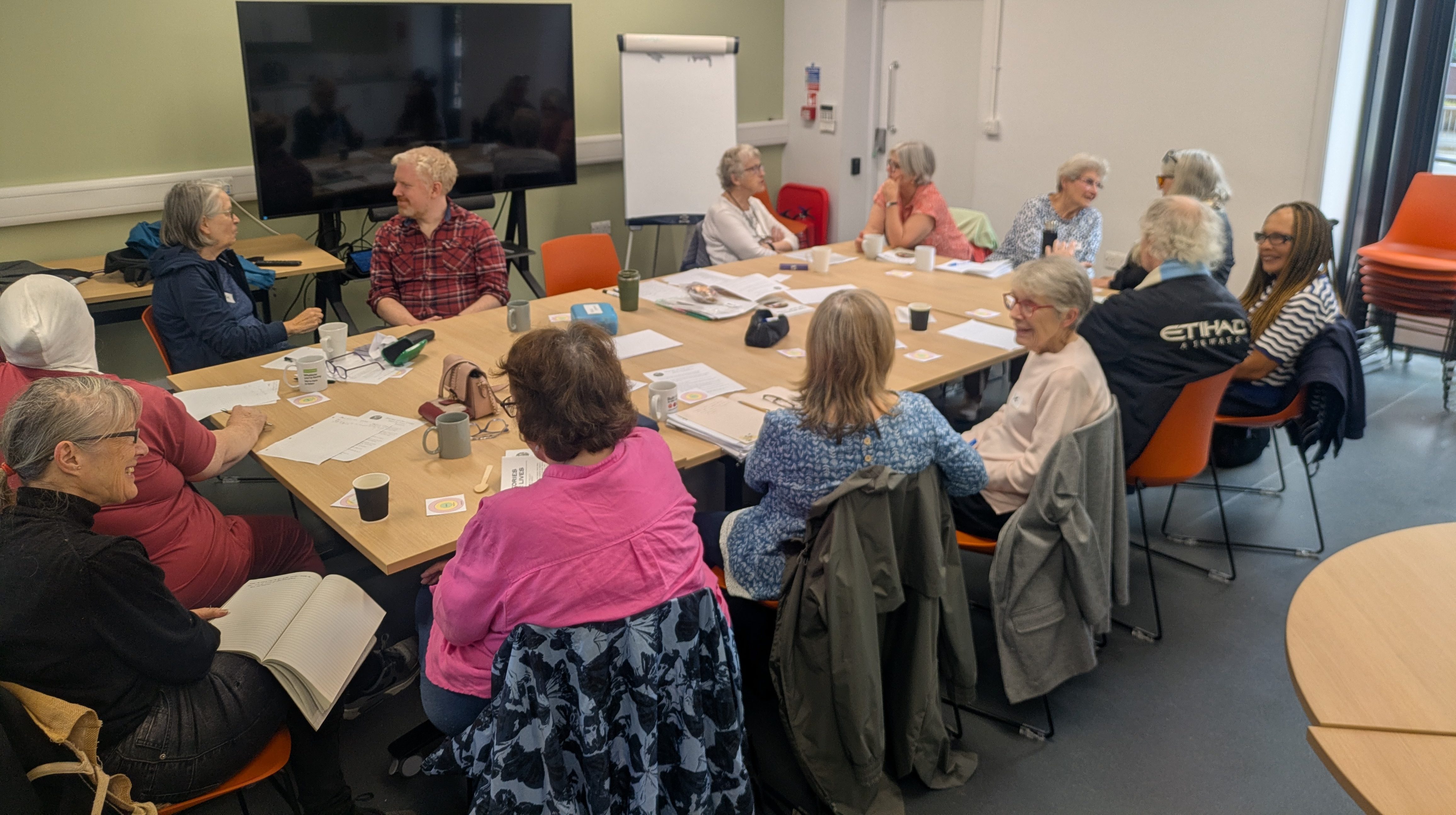 Image description: A group of around fourteen adults sit together around tables in a bright community room, chatting and smiling over cups of tea, papers and activity materials.
