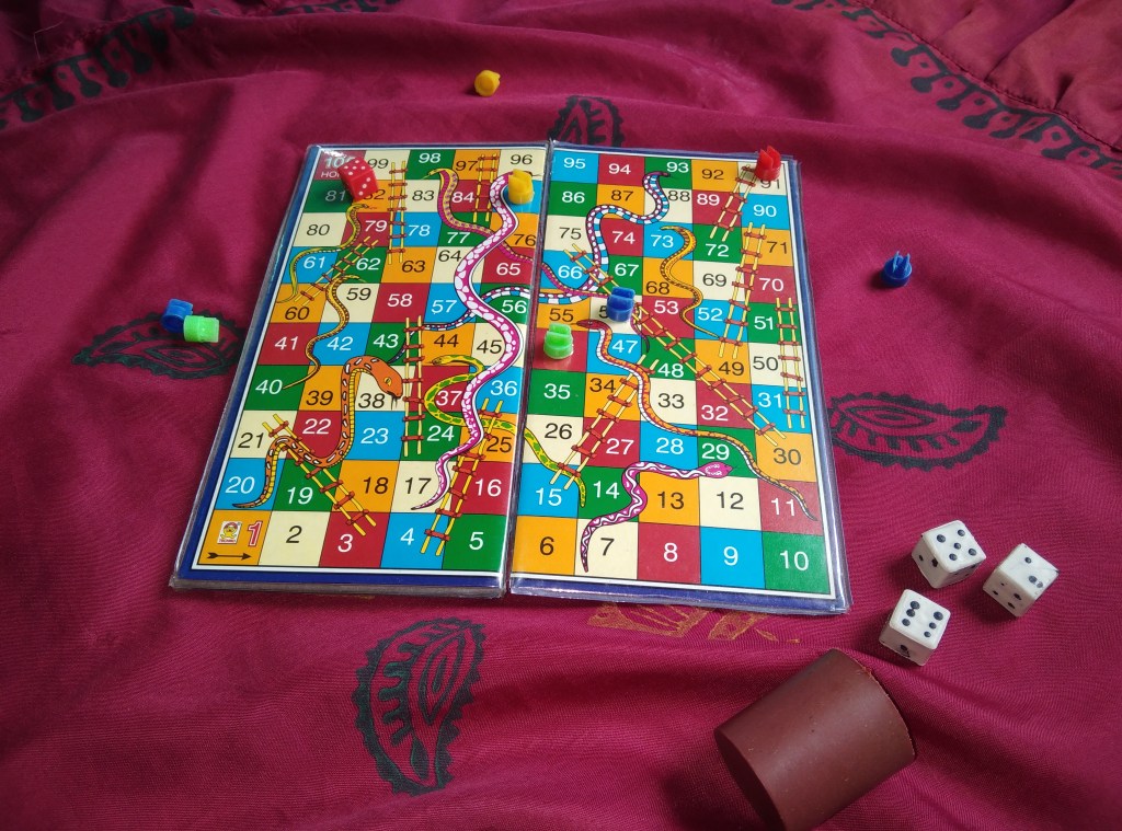 Image description: A Snakes and Ladders board game numbered from 1 to 100, with nine colourful snakes and ten ladders, all of different lengths, photographed on a red silky tablecloth, with three dice and playing pieces for four people. 