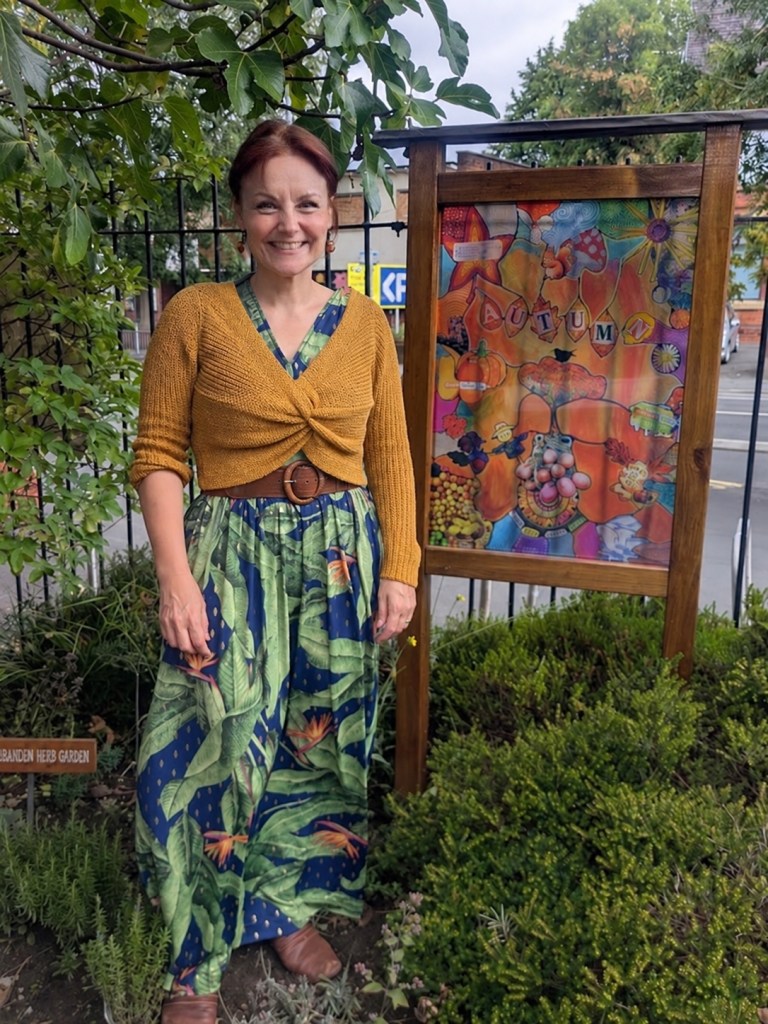 Image description: A smiling woman stands beside a wooden-framed community artwork in a leafy garden. The artwork is filled with bright autumn colours, leaves, fruit, seasonal images and the word “AUTUMN” across the centre.