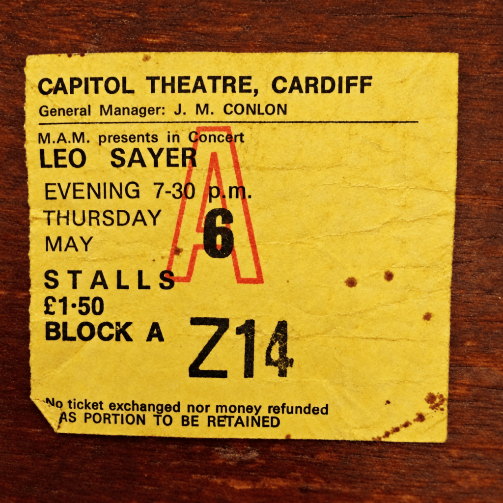 An original, aged yellow concert ticket for Leo Sayer at the Capitol Theatre, Cardiff, on Thursday 6 May at 7.30pm. The ticket is for the stalls, Block A, seat Z14, priced at £1.50. A large red letter A is stamped across the centre. The ticket shows signs of age, with creases, worn edges and brown marks, and rests on a dark wooden surface.